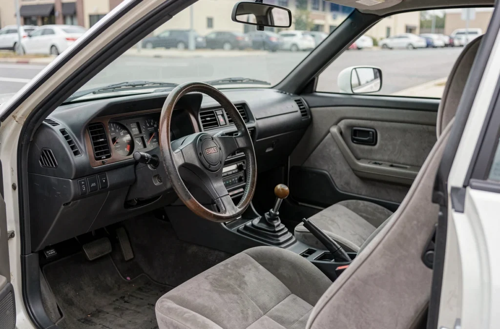 Intérieur authentique de la Toyota AE86 Trueno Corolla avec tableau de bord analogique, volant fin et sièges baquets en tissu usé, pris depuis la portière conducteur ouverte